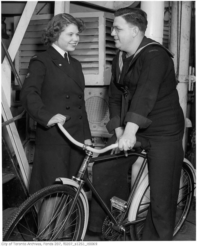 HMCS "Forest Hill" crew host WRENS stationed in Halifax from Forest Hill Village, and other parts of Toronto at a dinner on the ship : Coder James Cramer (1300 Lansdowne Ave.) shows WREN Ruth Arnsby (9 Montclair) one of the bicycles sent by the Village of Forest Hill to the ship 1940-45 - Vintage Bicycle Photographs