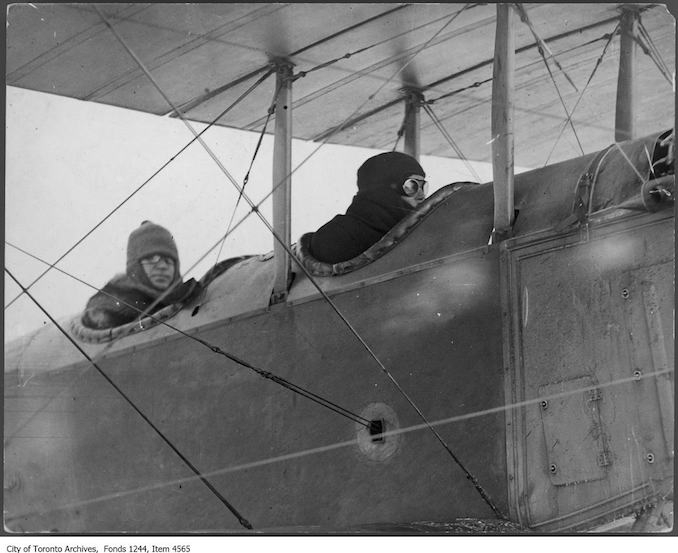 Aviator Bert Acosta prepares to take William James Sr. up in airplane, Leaside. - 1916 - photograph taken just before William James Sr. took the first movies from the air in Canada. Other subjects include F.G. Ericson (in checkered cap) and head of construction Sir Frank Baillie (right). - photograph . Baillie was the head of airplane construction. The pilot is Bert Acosta. The plane is a Jenny JN4.