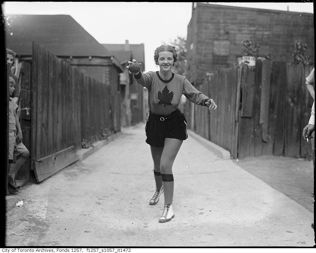 Billie Hallam, Miss Toronto 1937, wearing Toronto Maple Leafs Baseball Club outfit, striking baseball pose in alley 1937