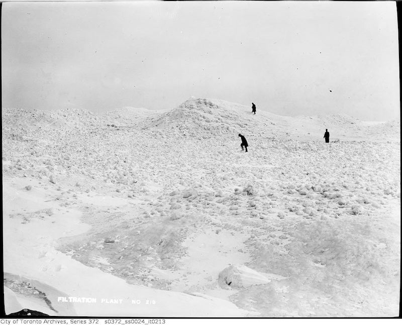 1911 - Island lake shore in winter showing snow and ice buildup, figures in distance