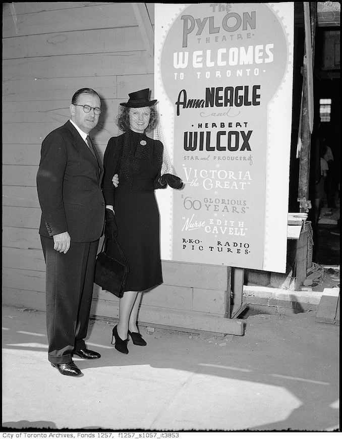 1939 - Anna Neagle and Herbert Wilcox, by sign advertising their movies