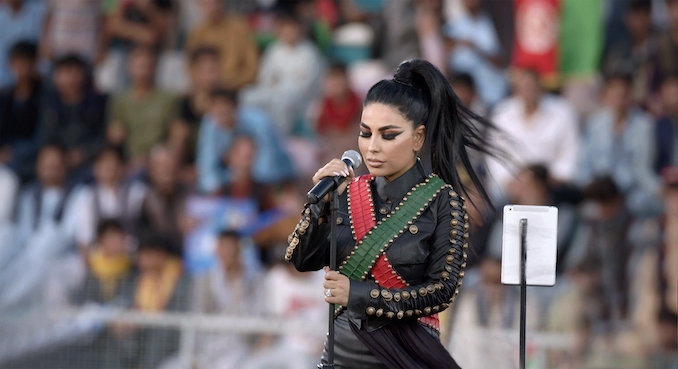 Aryana Sayeed singing at a microphone in conert in what looks to be an outdoor event space